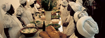 Women in  beauatiful white dresses and scarves stand at a table full of food they have prepared