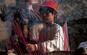 a young Thai boy, with a red hat, burns incense at a temple